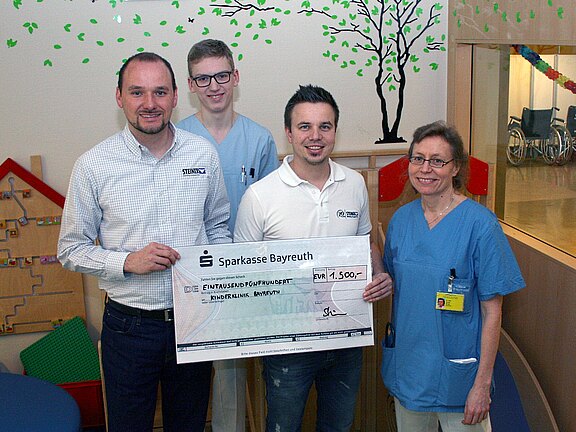 Vier Erwachsene, zwei in Klinikbekleidung, posieren in einem Krankenhaus und halten einen übergroßen Spendenscheck der Sparkasse Bayreuth über 1.500 Euro. Gruppenfoto zur Spendenübergabe.