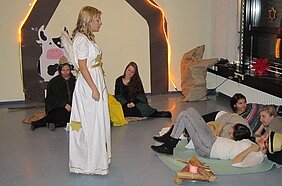 Eine Gruppe von Menschen sitzt auf dem Boden in einem Raum. Eine Person in einem weißen Kleid steht im Vordergrund und spricht. Im Hintergrund sind einige dekorative Elemente zu sehen. Die anderen Zuhörer scheinen aufmerksam zu sein und verfolgen die Darbietung.
