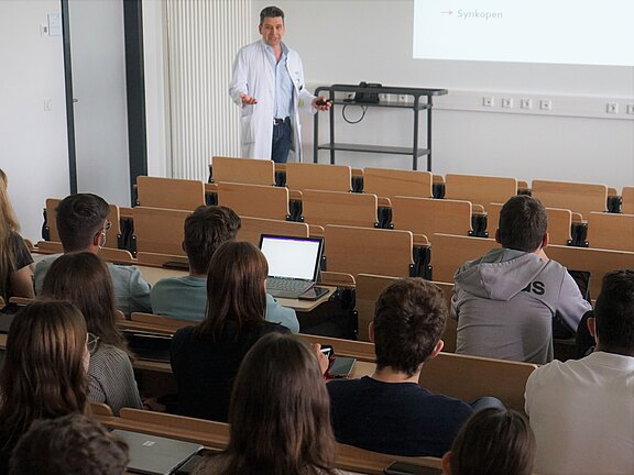 Ein Dozent im weißen Kittel hält eine Vorlesung in einem Hörsaal. Vor ihm sitzen Studierende mit Laptops. Auf der Leinwand ist eine Präsentationsfolie zum medizinischen Thema „Synkopen“ zu sehen.