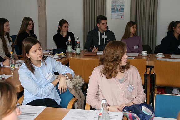 Mehrere junge Erwachsene sitzen in einem Seminarraum an Tischen, hören einer Präsentation zu und schauen nach vorn; vor ihnen liegen Unterlagen. Die Szene wirkt wie ein Workshop oder eine Schulung.