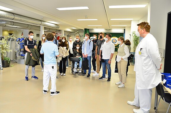 Gruppe junger Erwachsener in einem Klinikraum hört einer Pflegekraft zu; ein Arzt im weißen Kittel steht daneben. Alle tragen Masken. Im Hintergrund medizinische Geräte. Die Szene wirkt wie eine Führung oder Einweisung; eine Person sitzt im Rollstuhl.