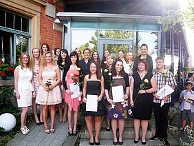 Eine Gruppe von jungen Frauen und einem Mann steht auf einer Treppe vor einem Gebäude und hält Dokumente sowie Blumen. Alle zeigen lächelnd ihre Abschlusszertifikate. Die Atmosphäre ist feierlich und fröhlich, mit Sonnenlicht, das auf die Szene strahlt.