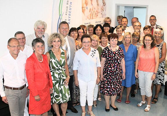 Gruppenfoto von etwa 30 Erwachsenen in einem hellen Raum. Sie stehen dicht zusammen, lächeln in die Kamera und wirken wie Kolleginnen und Kollegen bei einer offiziellen Veranstaltung oder Ehrung. Unterschiedliche Kleidung, teils sommerlich, vermittelt eine freundliche, festliche Stimmung.