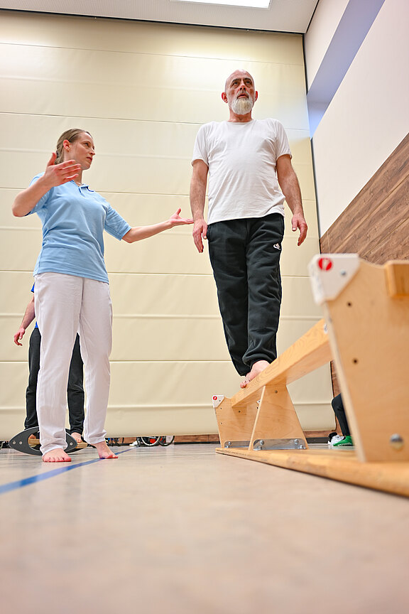 Ein älterer Mann balanciert barfuß auf einer niedrigen Holzstange in einer Turnhalle. Eine Therapeutin neben ihm gibt Anweisungen und sichert ihn. Die Szene zeigt Gleichgewichtstraining beziehungsweise Physiotherapie.