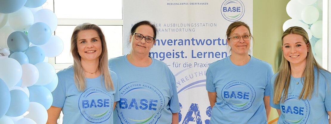 Vier Personen in hellblauen BASE-T-Shirts posieren vor einem Banner des Klinikums Bayreuth. Die Szene wirbt für die Ausbildungsstation BASE; freundliches Gruppenfoto bei einer Informations- oder Eröffnungsveranstaltung.