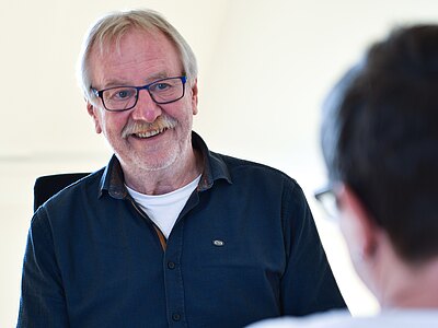 Älterer Mann mit Brille lächelt und spricht mit einer unscharfen Person im Vordergrund. Die Szene vermittelt ein freundliches Gespräch oder eine Beratung in heller Umgebung.