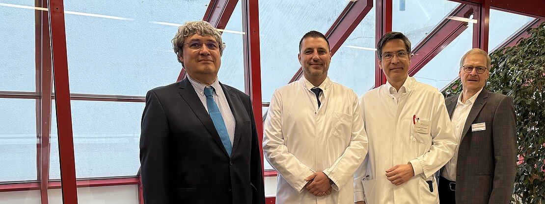Vier Männer stehen in einem hellen Gebäudeflur; zwei tragen weiße Arztkittel, zwei Businesskleidung. Sie posieren nebeneinander und blicken in die Kamera. Große Fenster mit roten Rahmen im Hintergrund. Szene wirkt professionell, vermutlich Krankenhaus oder Klinik.