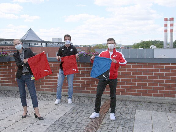 Drei Personen mit Gesichtsmasken stehen im Freien auf Abstand. Sie halten Puma-Turnbeutel hoch: zwei rote, einer blau sowie ein schwarzer. Präsentation von Sportbeuteln.