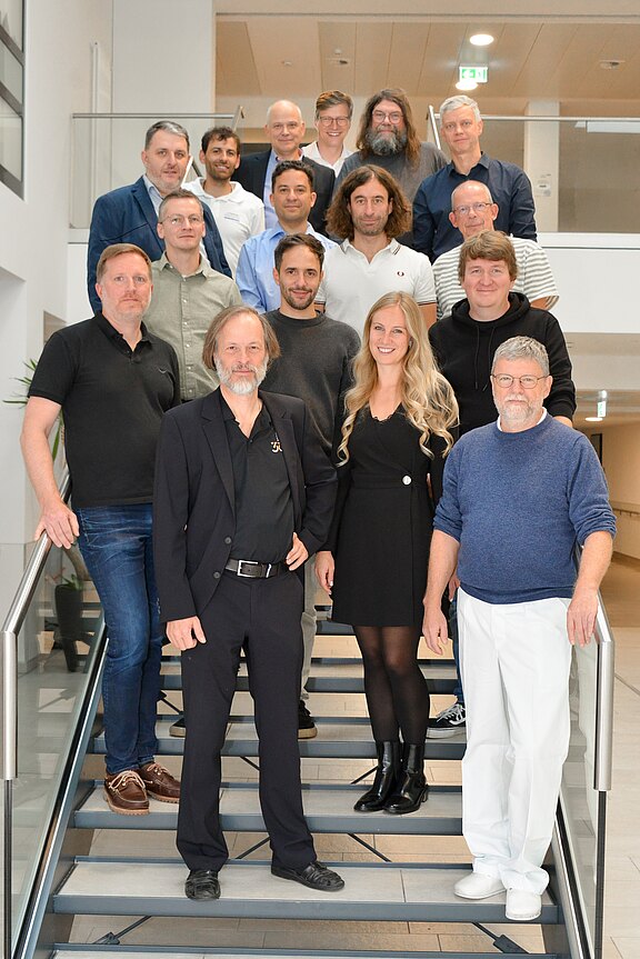 Gruppenfoto: Rund 16 Erwachsene stehen in mehreren Reihen auf einer Treppe in einem hellen, modernen Gebäude und blicken in die Kamera. Mischung aus formeller und legerer Kleidung.