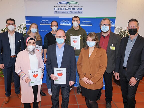 Gruppenfoto in einem Krankenhaus: zehn Personen mit Masken stehen vor einem Banner „Klinikum Bayreuth GmbH“. Drei Personen vorn halten Urkunden mit rotem Herz. Es wirkt wie eine Ehrung oder Dankesveranstaltung, vermutlich für Engagement im Gesundheitsbereich.