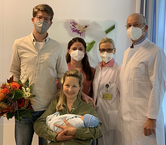 Eine Mutter hält ein neugeborenes Baby in einer blauen Decke und lächelt. Neben ihr stehen ein Mann mit einem Blumenstrauß, eine Krankenschwester und zwei Ärzte, alle tragen Schutzmasken. Der Hintergrund zeigt unscharfe Pflanzen.