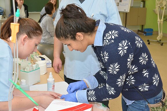 Zwei Frauen arbeiten in einem Schulungsraum zusammen. Eine Frau trägt Handschuhe und untersucht die Hand der anderen. Auf dem Tisch sind medizinische Materialien und Flaschen sichtbar. Im Hintergrund sind weitere Personen zu sehen, die ebenfalls an einem Lernprozess beteiligt sind.