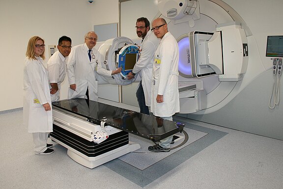 Fünf Personen in weißen Kitteln stehen in einem Behandlungsraum neben einem großen Linearbeschleuniger für Strahlentherapie. Sie justieren ein Gerät am Strahler; davor befindet sich die flache Behandlungsliege. Moderne, klinische Umgebung.