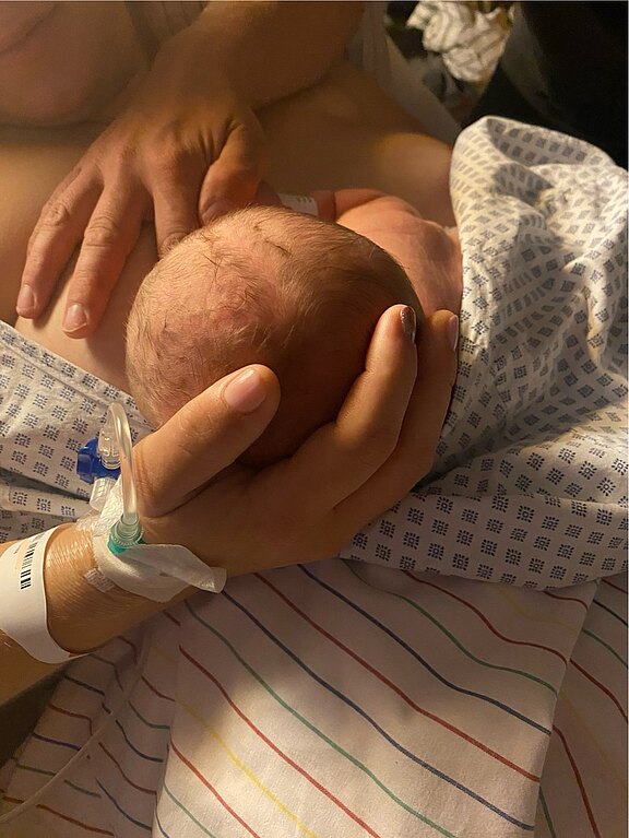 Neugeborenes liegt auf der Brust eines Elternteils im Krankenhaus, in Tücher gewickelt. Hände stützen den Kopf; ein Infusionszugang am Handgelenk ist sichtbar. Szene zeigt erstes Haut-an-Haut-Kuscheln nach der Geburt.