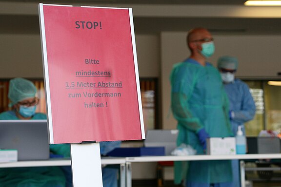 Ein Stoppschild fordert auf, mindestens 1,5 Meter Abstand zum Vordermann zu halten. Im Hintergrund sind medizinisches Personal in Schutzkleidung und mit Masken zu sehen, das für die Einhaltung der Abstandsregelungen sorgt.