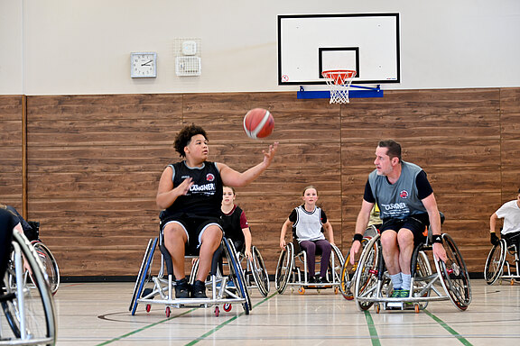 Mehrere Personen spielen Rollstuhlbasketball in einer Turnhalle. Im Vordergrund passt ein Spieler den Ball, ein anderer fährt daneben; im Hintergrund das Korbbrett. Fokus liegt auf dynamischem Spiel und Teamarbeit.