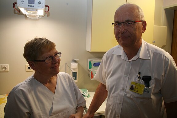 Zwei medizinische Fachkräfte in weißer Kleidung stehen in einem Behandlungsraum und unterhalten sich; eine Person blickt lächelnd zur anderen. Medizinische Geräte und Schränke im Hintergrund.