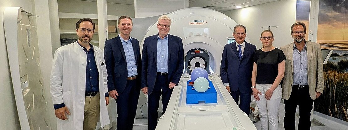 Sechs Personen stehen zur Kamera. Sie befinden sich in einem modernen medizinischen Raum neben einem MRT-Gerät von Siemens. Auf der Liege des Geräts liegen mehrere farbige Luftballons. Der Raum wirkt klinisch und gut beleuchtet, mit neutraler Farbgestaltung.