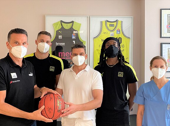 Eine Gruppe von vier Personen steht zusammen, trägt Masken und hält einen Basketball. Im Hintergrund sind zwei signierte Trikots zu sehen. Die Personen sind sportlich gekleidet und stehen in einem gut beleuchteten Raum.