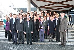 Eine Gruppe von etwa 40 Personen steht auf einer Treppe vor einem modernen Gebäude. Die Teilnehmer sind in formeller Kleidung und lächeln. Die Atmosphäre wirkt freundlich und einladend, was auf eine Veranstaltung oder ein Treffen hinweist.