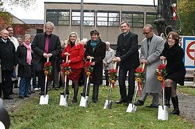 Eine Gruppe von fünf Personen steht mit Schaufeln in der Hand in einer Gartenlandschaft, bereit, einen Spatenstich zu vollziehen. Sie tragen festliche Kleidung und halten Blumensträuße. Im Hintergrund sind Zuschauer und ein Gebäude zu sehen, was auf eine öffentliche Veranstaltung hinweist.