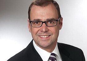 Ein lächelnder Mann mit Brille und einem Anzug. Er hat kurze, braune Haare und trägt eine gestreifte Krawatte. Der Hintergrund ist verschwommen und hell.