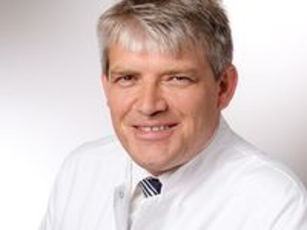 Ein lächelnder Arzt trägt einen weißen Kittel und eine Krawatte. Der Hintergrund ist hell und neutral. Die Person hat kurzes, graues Haar und blickt direkt in die Kamera.