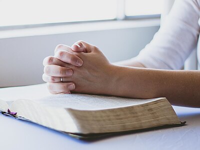 Verschränkte Hände ruhen auf einer aufgeschlagenen Bibel; die Person betet oder meditiert in ruhiger, lichtdurchfluteter Umgebung.