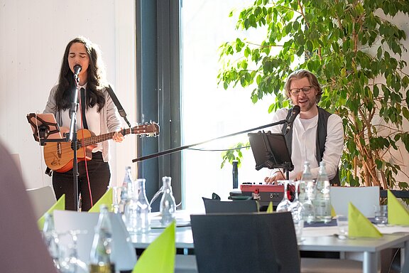 Zwei Musiker*innen treten in einem hellen Raum auf: links singt eine Person und spielt eine kleine Gitarre, rechts singt eine Person am Keyboard mit Mikrofon.