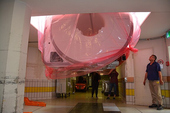 Ein großer medizinischer Scanner hängt von der Decke in einem Krankenhausflur. Er ist mit einer transparenten Plastikhülle geschützt. Zwei Personen stehen darunter und schauen auf das Gerät, während ein weiterer Mitarbeiter im Hintergrund auf einen anderen Wagen schaut.