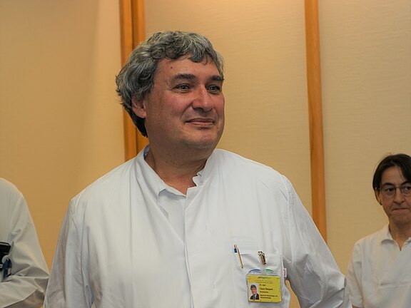 Ein Arzt mit grauen Haaren trägt einen weißen Kittel und lächelt. Er steht in einem Raum, in dem eine weitere Person ebenfalls in einem weißen Kittel zu sehen ist. Die Atmosphäre wirkt freundlich und professionell.