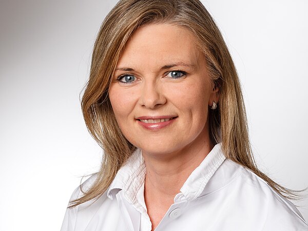 Porträt einer lächelnden Person mit schulterlangem blondem Haar in weißem Kittel vor neutralem hellgrauem Hintergrund.