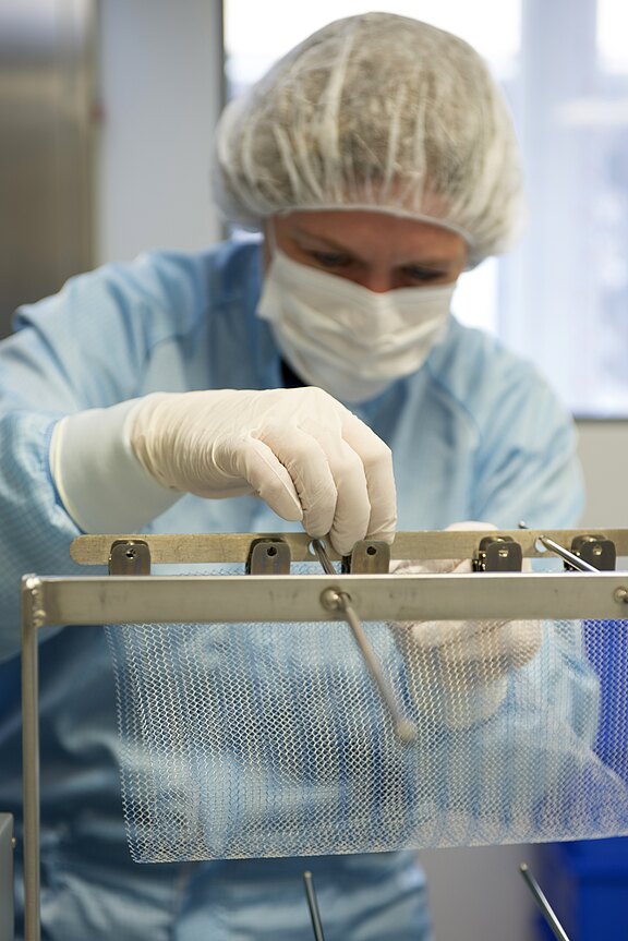 Eine Person in Schutzkleidung und Handschuhen arbeitet vorsichtig mit medizinischen Instrumenten an einem Metallgitter. Der Fokus liegt auf ihrer Hand, die gerade einen Gegenstand greift. Im Hintergrund sind verschwommene medizinische Geräte sichtbar.