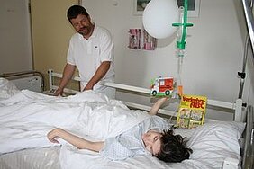 Ein Pfleger steht an einem Krankenhausbett, in dem ein Kind mit einem Verband schläft. Das Kind hält ein Spielzeug-Lastwagen und ein Buch in der Hand. Im Hintergrund ist ein Tropf zu sehen, der über dem Bett hängt.