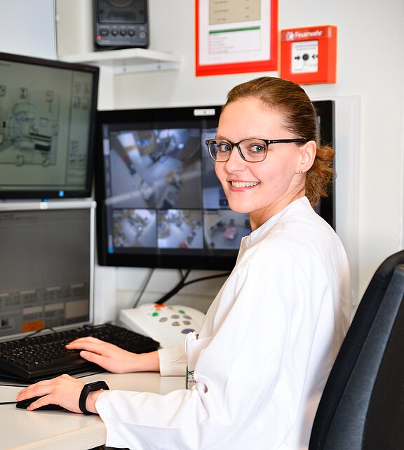 Mitarbeiterin in weißem Kittel sitzt am Kontrollplatz mit mehreren Monitoren, bedient Maus und Tastatur und lächelt zur Kamera. Auf den Bildschirmen sind mehrere Kameraansichten eines Behandlungsraums zu sehen. Die Szene zeigt Überwachung und Steuerung in einem medizinischen Umfeld.