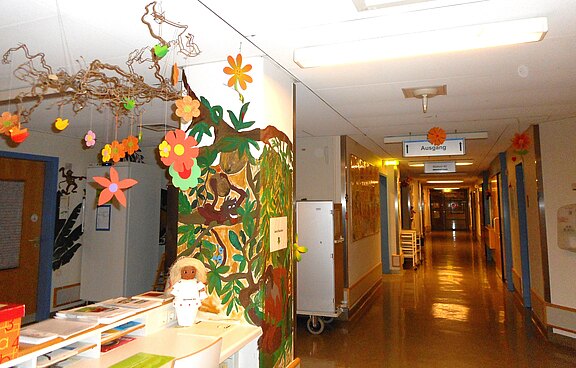 Krankenhausflur, farbenfroh dekoriert mit Papierblumen und Dschungel-Malerei; freundliche Atmosphäre, Wegweiser „Ausgang“ sichtbar.