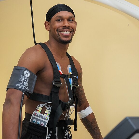 Muskulöser Mann beim medizinischen Belastungstest: Oberkörper frei, EKG-Elektroden auf der Brust, Gurte und Messgeräte am Bauch, Blutdruckmanschette am Arm. Er steht auf einem Laufband und lächelt in die Kamera.