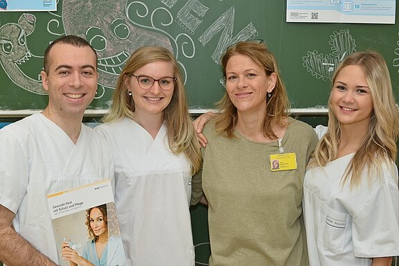 Vier Personen in medizinischer Kleidung stehen zusammen vor einer Tafel. Der Mann und zwei Frauen tragen weiße Kittel, die vierte Frau trägt ein graues Oberteil. Eine der Frauen hält ein Heft mit Informationen zur Hautpflege. Alle lächeln freundlich in die Kamera.