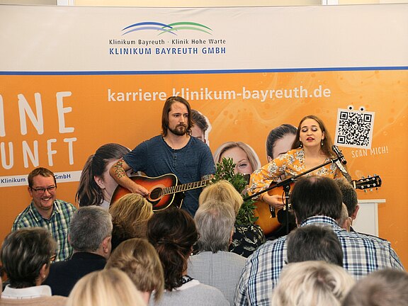 Zwei Musiker mit Akustikgitarren treten vor einem sitzenden Publikum auf. Die Sängerin singt ins Mikrofon, der Mann begleitet auf der Gitarre. Im Hintergrund hängt ein orangefarbenes Werbebanner.