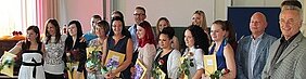 Eine Gruppe von Absolventen steht auf einer Bühne und hält ihre Zertifikate in den Händen. Einige von ihnen tragen Blumen. Es gibt auch einige Personen im Hintergrund, die die Absolventen unterstützen. Die Stimmung wirkt feierlich und erfreulich.