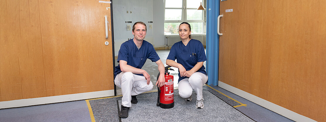 Zwei Personen in blauen Pflegekleidern knien in einem Krankenhausflur und präsentieren einen roten Feuerlöscher zwischen sich. Geöffnete Doppeltüren deuten auf eine Brandschutzübung hin.