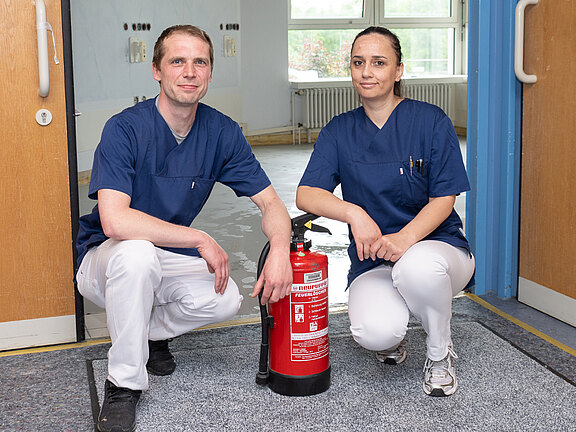 Zwei Personen in blauen Pflegekleidern knien in einem Krankenhausflur und präsentieren einen roten Feuerlöscher zwischen sich. Geöffnete Doppeltüren deuten auf eine Brandschutzübung hin.