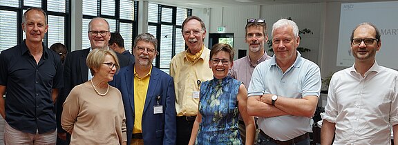 Gruppe von neun Erwachsenen steht in einem hellen Konferenzraum, lächelt in die Kamera. Einige tragen Namensschilder; Kleidung ist business-casual. Stimmung freundlich, wirkt wie ein Workshop oder Netzwerktreffen.