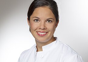 Porträt einer lächelnden Person in weißem Kittel vor neutralem Hintergrund; Kopf-und-Schulter-Aufnahme, professionell und freundlich.