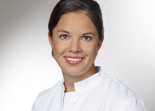 Porträt einer lächelnden Person in weißem Kittel vor neutralem Hintergrund; Kopf-und-Schulter-Aufnahme, professionell und freundlich.