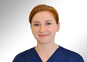 Station 14 Porträtfoto einer lächelnden Frau in blauer medizinischer Arbeitskleidung vor hellem, neutralem Hintergrund; Schulteransicht, Blick in die Kamera.