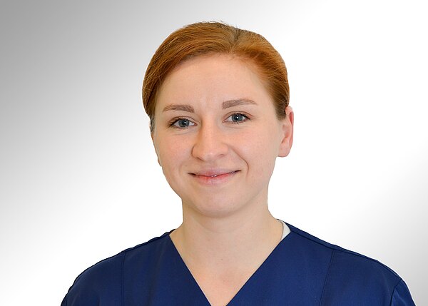 Porträtfoto einer lächelnden Frau in blauer medizinischer Arbeitskleidung vor hellem, neutralem Hintergrund; Schulteransicht, Blick in die Kamera.