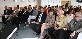 Eine große Gruppe von Menschen sitzt in einem Raum. Einige Teilnehmer tragen formelle Kleidung. Es scheint sich um eine Veranstaltung oder eine Konferenz zu handeln, bei der die Anwesenden aufmerksam auf die Bühne schauen oder miteinander sprechen.