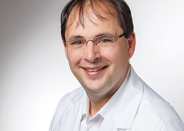 Porträtfoto: Ein lächelnder Mann mit Brille trägt einen weißen Kittel und blickt in die Kamera vor neutralem Hintergrund.