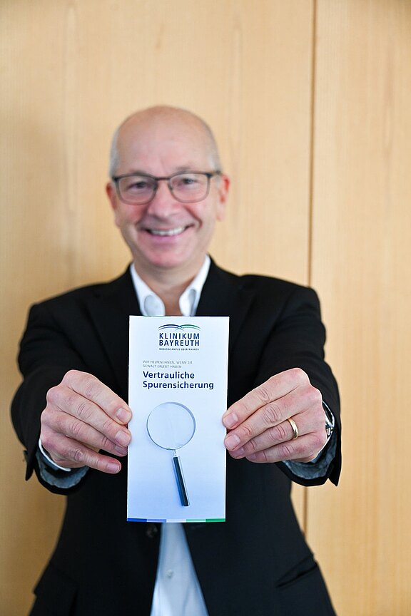 Ein lächelnder Mann im Anzug hält eine Broschüre in die Kamera. Auf der Vorderseite steht Klinikum Bayreuth und Vertrauliche Spurensicherung, dazu ein Symbol einer Lupe. Die Szene verweist auf ein Angebot zur vertraulichen Spurensicherung im Klinikum.